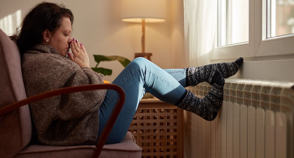 woman cold and bundled up in living room next to radiator or heater